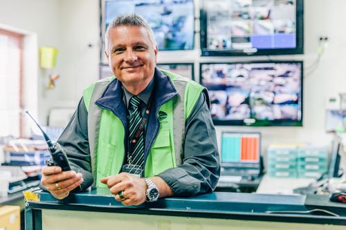 A smiling man in a high-visibility vest holding a radio, standing in an office with multiple surveillance monitors in the background.
