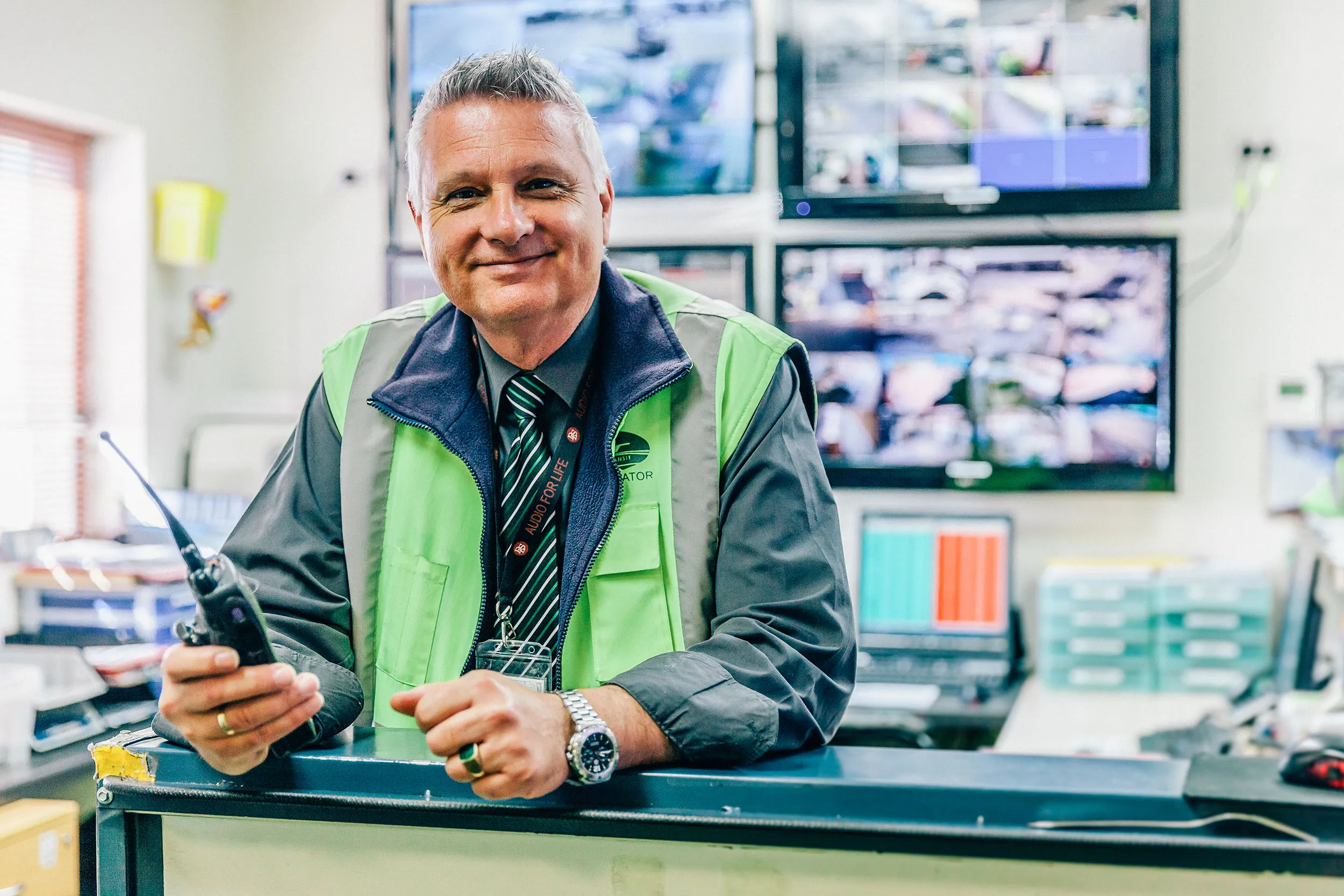 A smiling man in a high-visibility vest holding a radio, standing in an office with multiple surveillance monitors in the background.