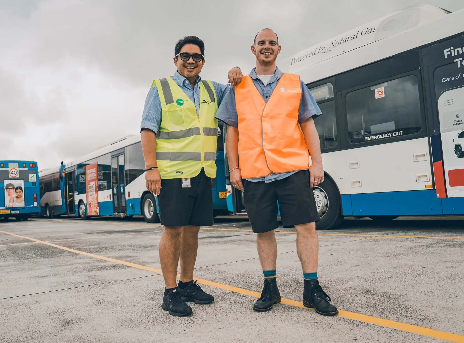 Two male bus drivers in high-visibility vests standing in front of parked buses, one with his arm around the other.