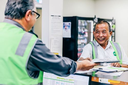 Two staff members in green safety vests interact at a counter in a workplace, one handing a document to the other who is smiling.