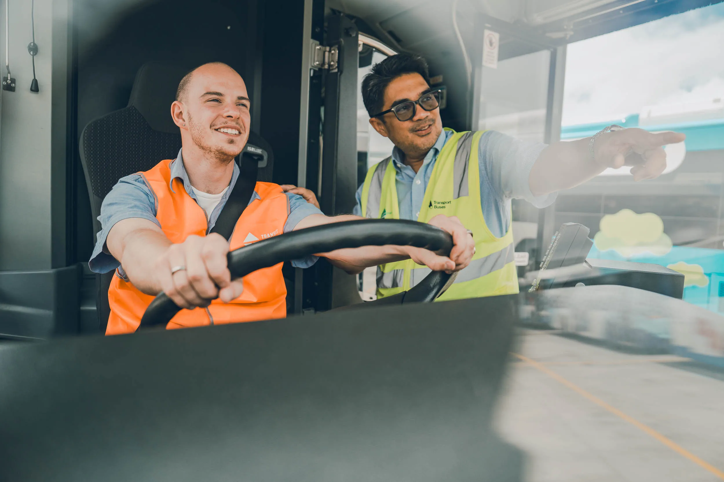 Two men, one driving and the other instructing, inside a bus, both wearing high-visibility vests.