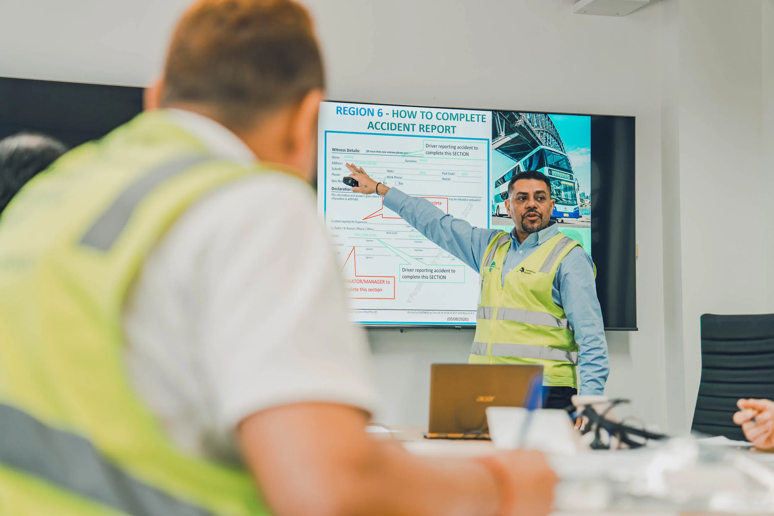 A man in a high-visibility vest pointing at a large screen displaying an "ACCIDENT REPORT" during a training session.