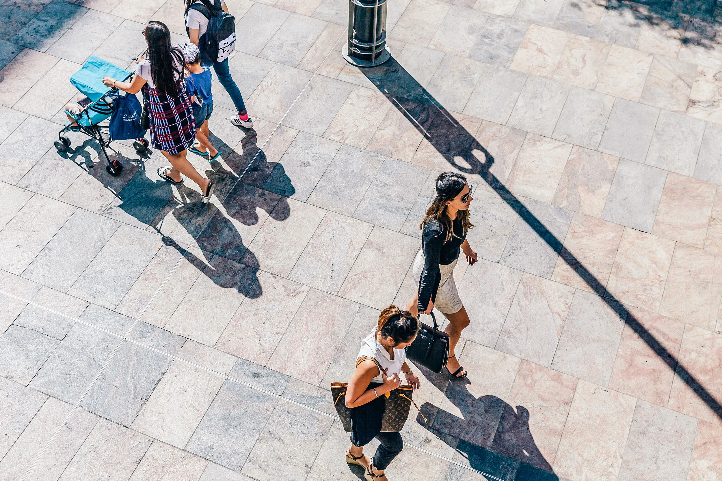 An overhead view of several people walking on a tiled outdoor plaza, casting long shadows.