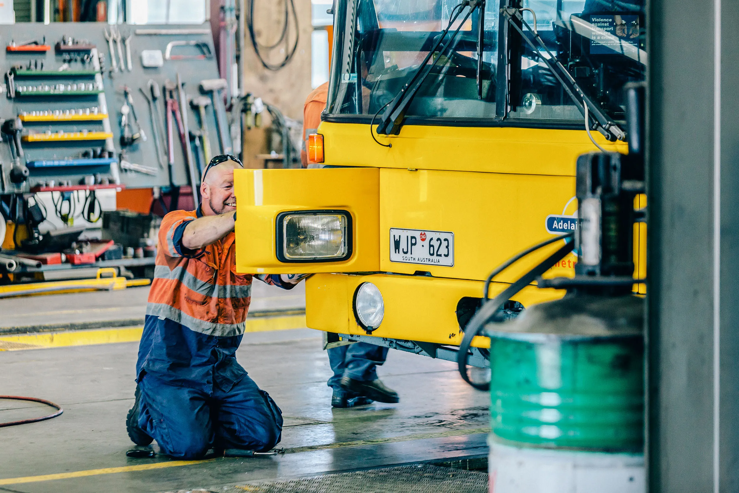 A male mechanic kneeling and working on the front of a yellow "Adelaide Metro" bus in a garage.