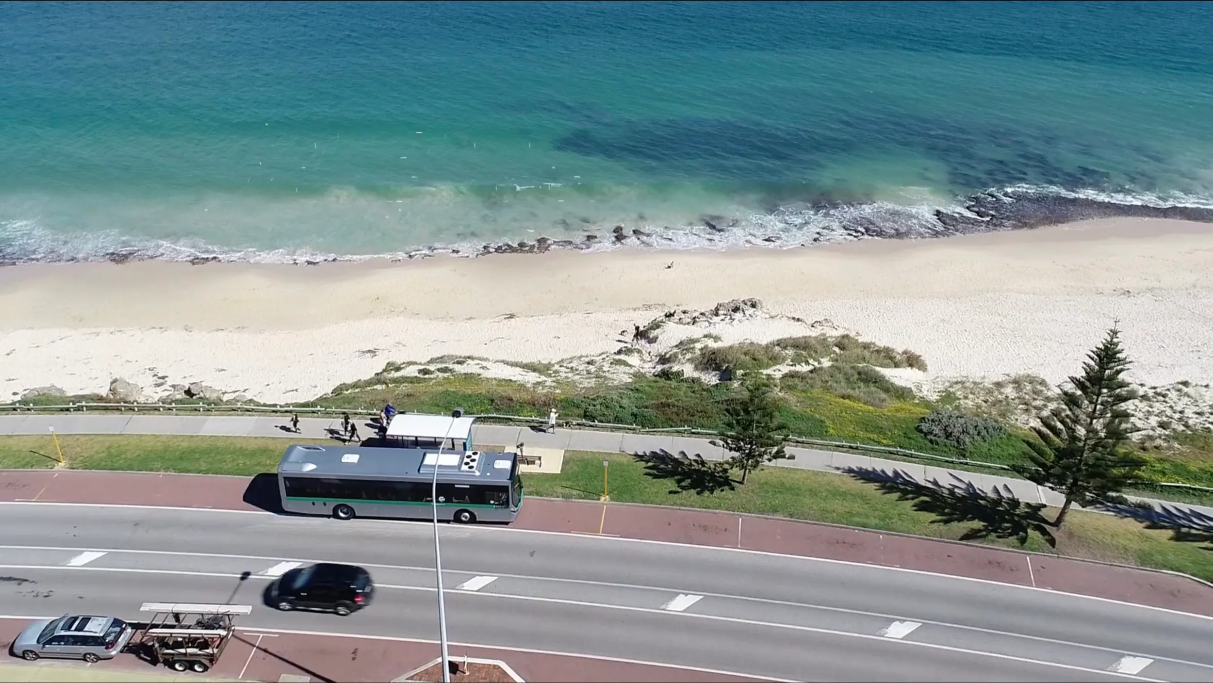 Aerial view of a bus on a coastal road next to a beach and turquoise ocean.