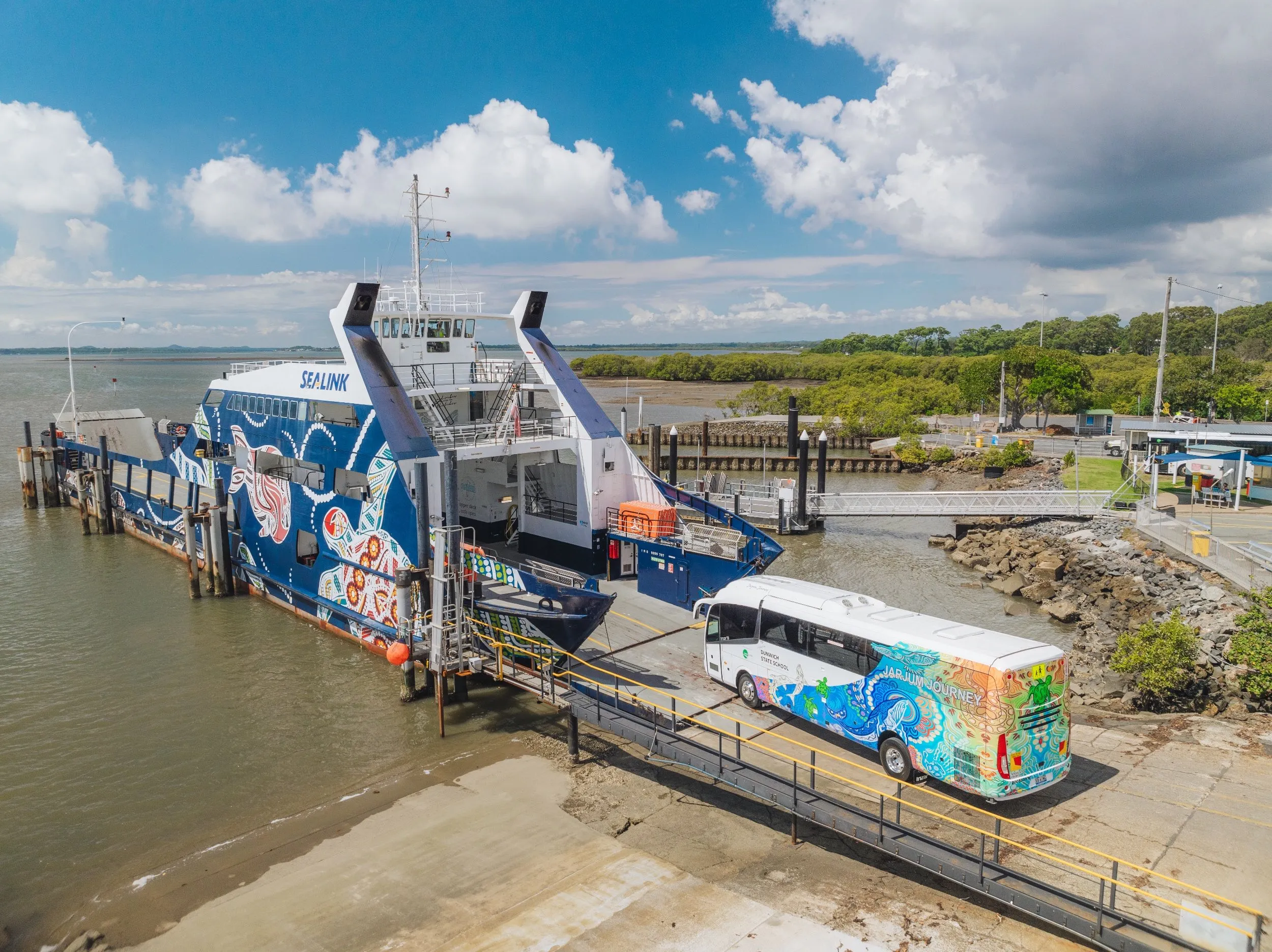 An aerial shot of a Sealink ferry, adorned with Indigenous artwork, docked next to a similarly decorated bus, with a body of water and lush green land in the background.