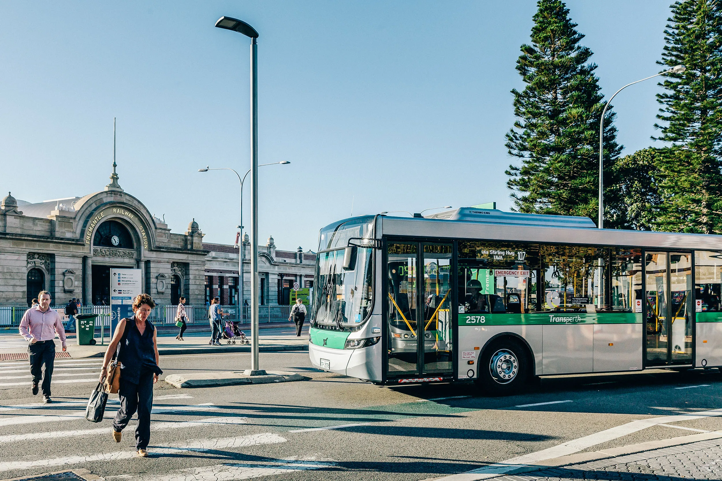 A "Transperth" bus at a crosswalk with people walking, and a historic building in the background.
