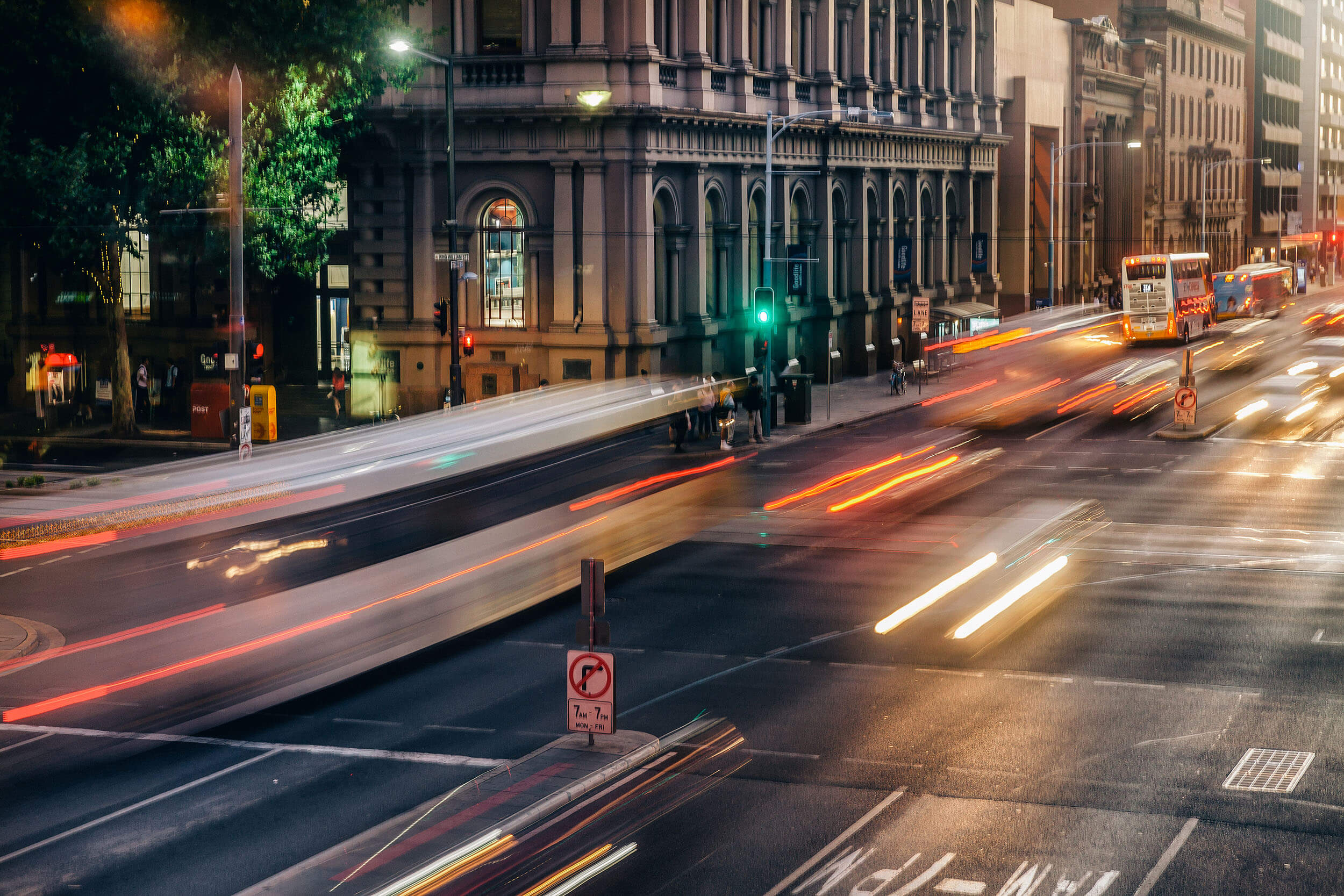 Long exposure shot of a city street at dusk with blurred light trails from moving vehicles and a building on the left.