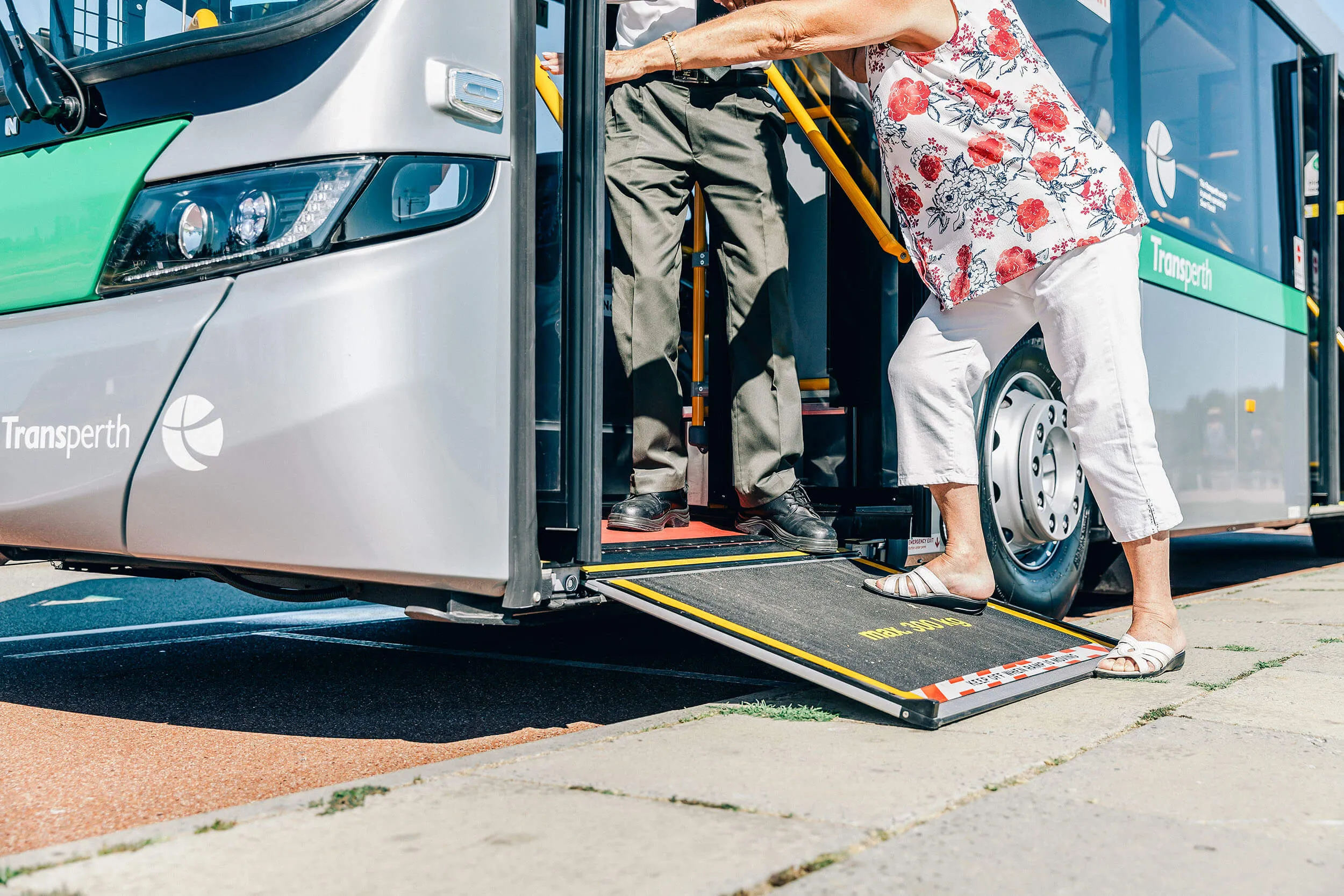 Two people boarding a Transperth bus using a deployed ramp, one in white pants and a floral top.