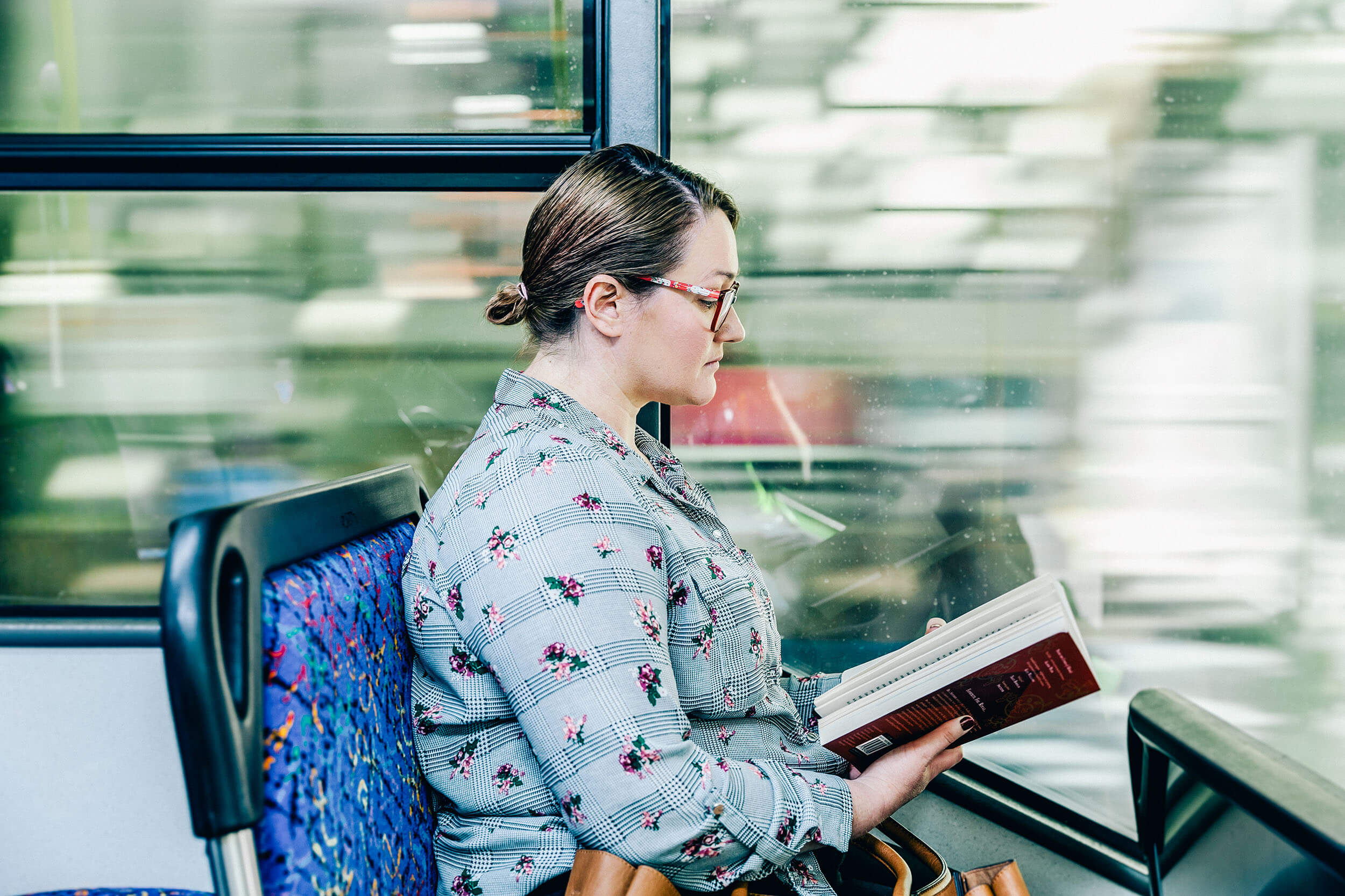 A woman with her hair in a bun and wearing glasses, reading a book while sitting on a bus.