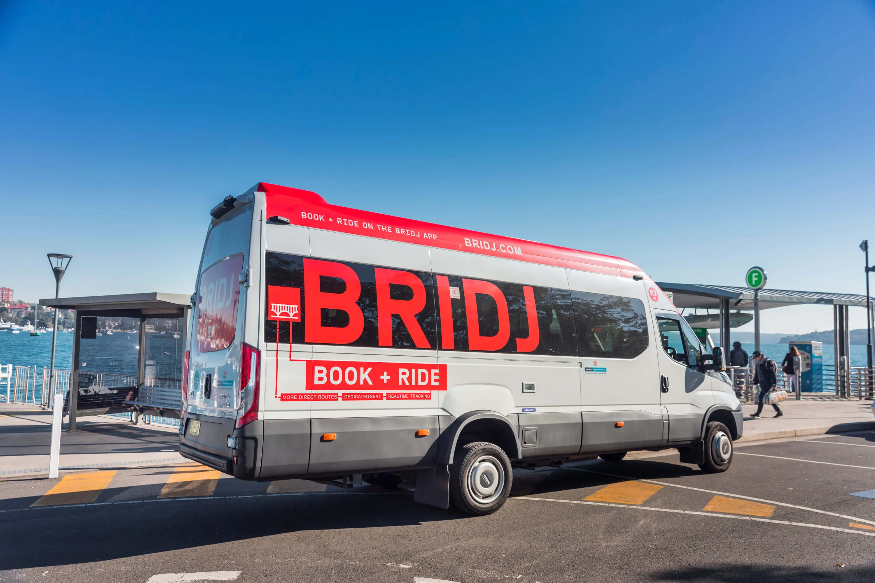 A white and grey "BRIDJ" van with red lettering, parked near a waterfront bus stop on a sunny day.