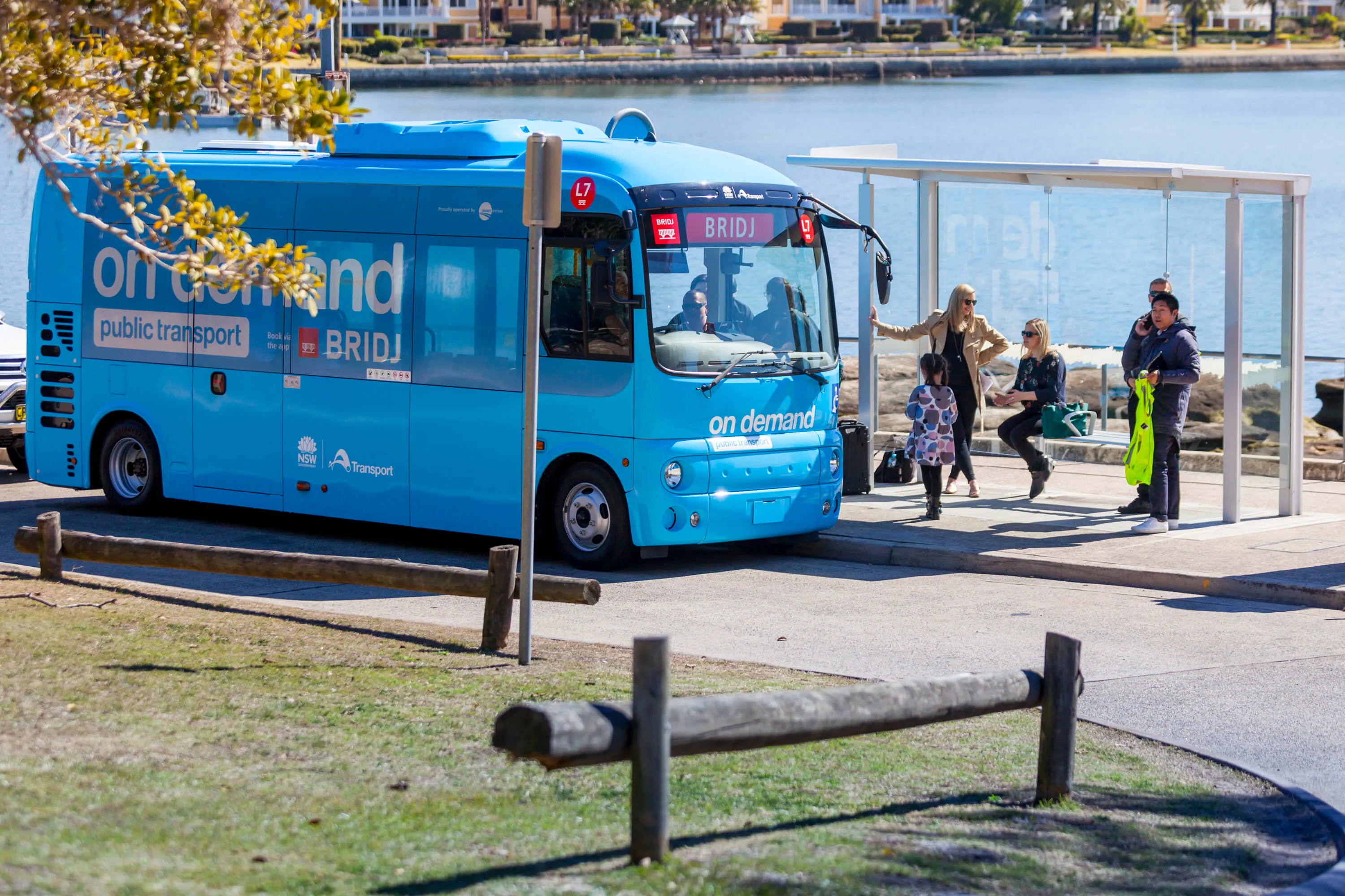 A blue "on demand" bus with "BRIDJ" on its front, stopped at a bus shelter with passengers waiting nearby, overlooking water.