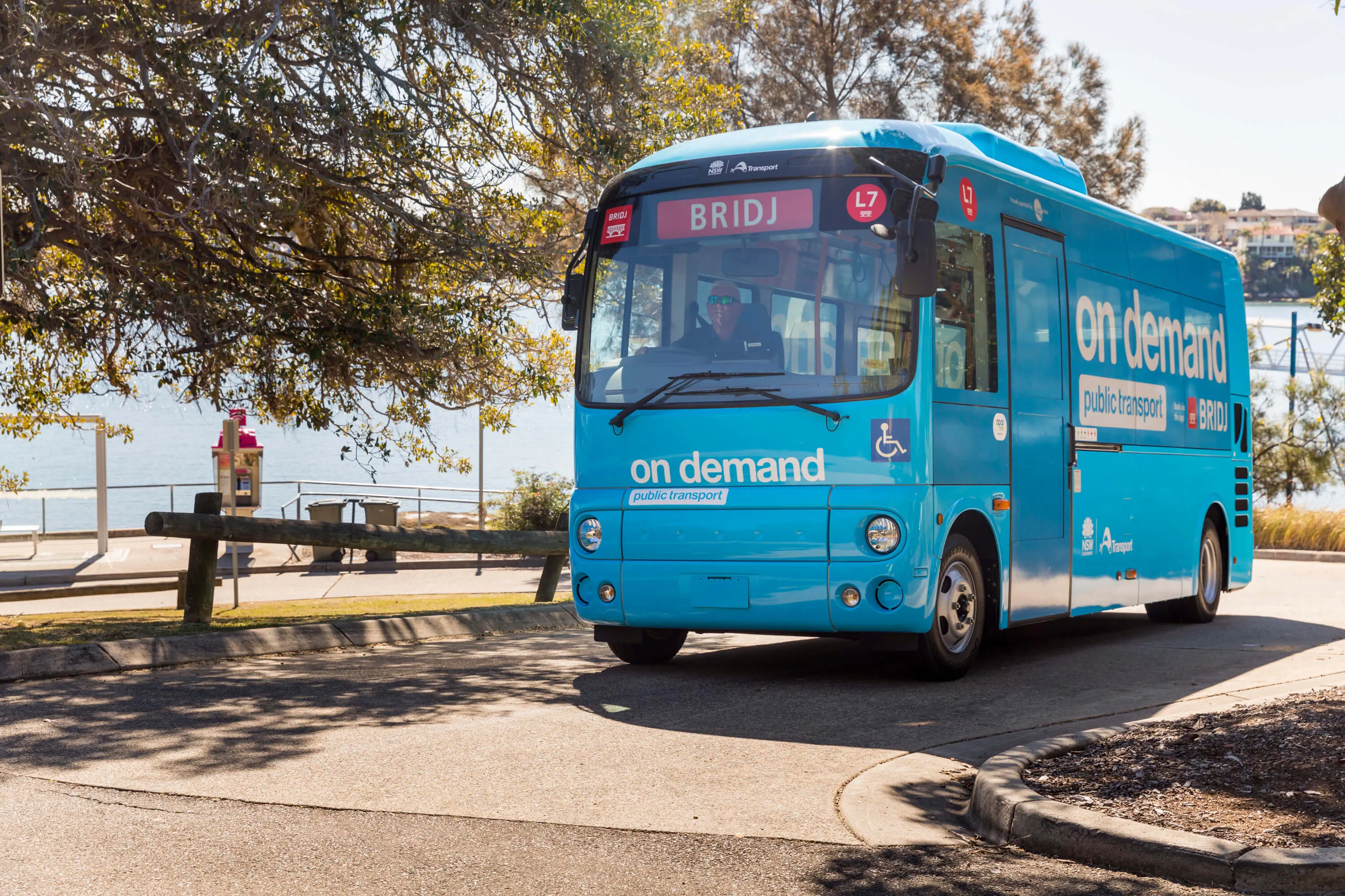 A blue "on demand" bus with "BRIDJ" on its front, parked near a waterfront.