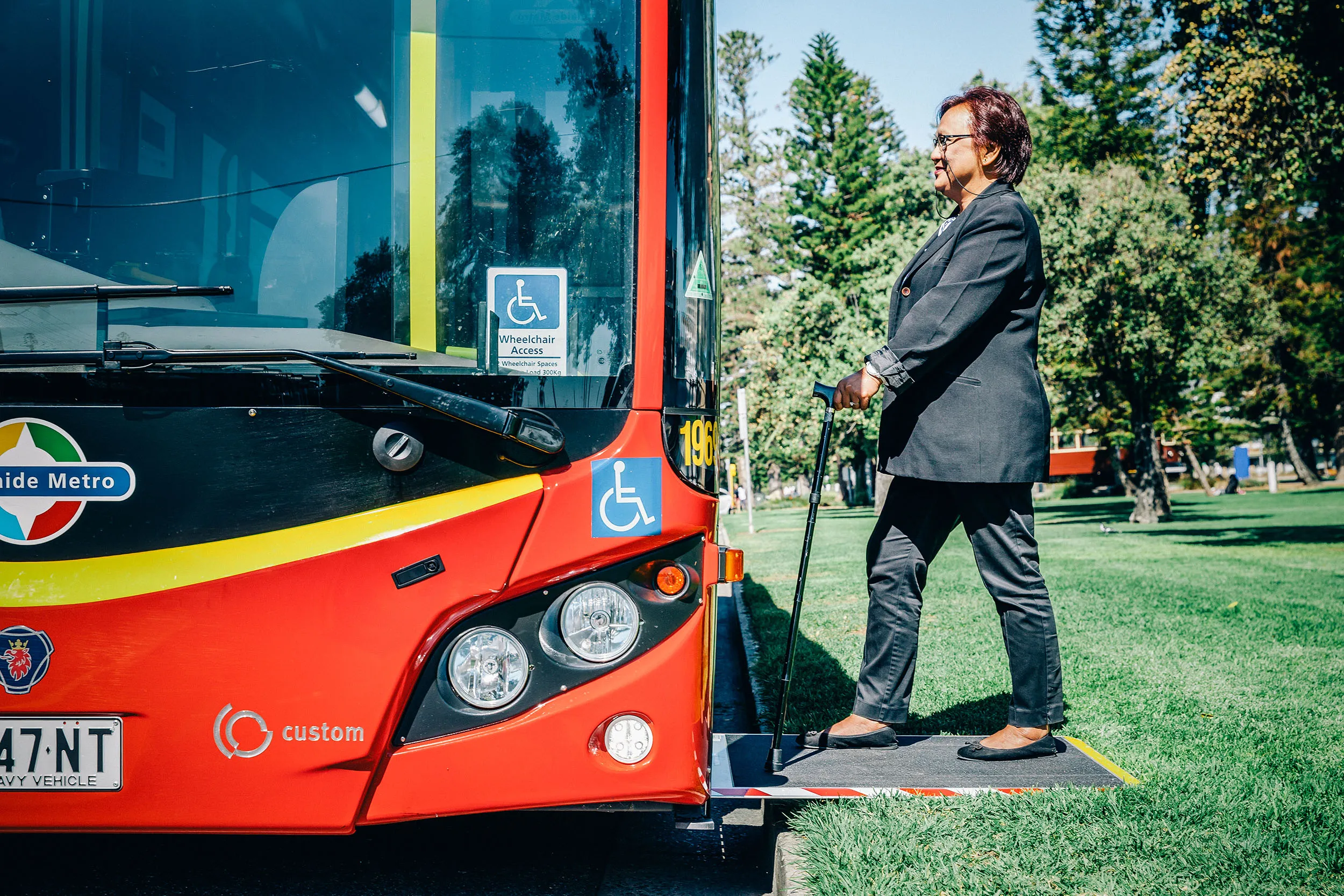 A woman with a walking stick standing on the lowered ramp of a red and black Adelaide Metro bus.