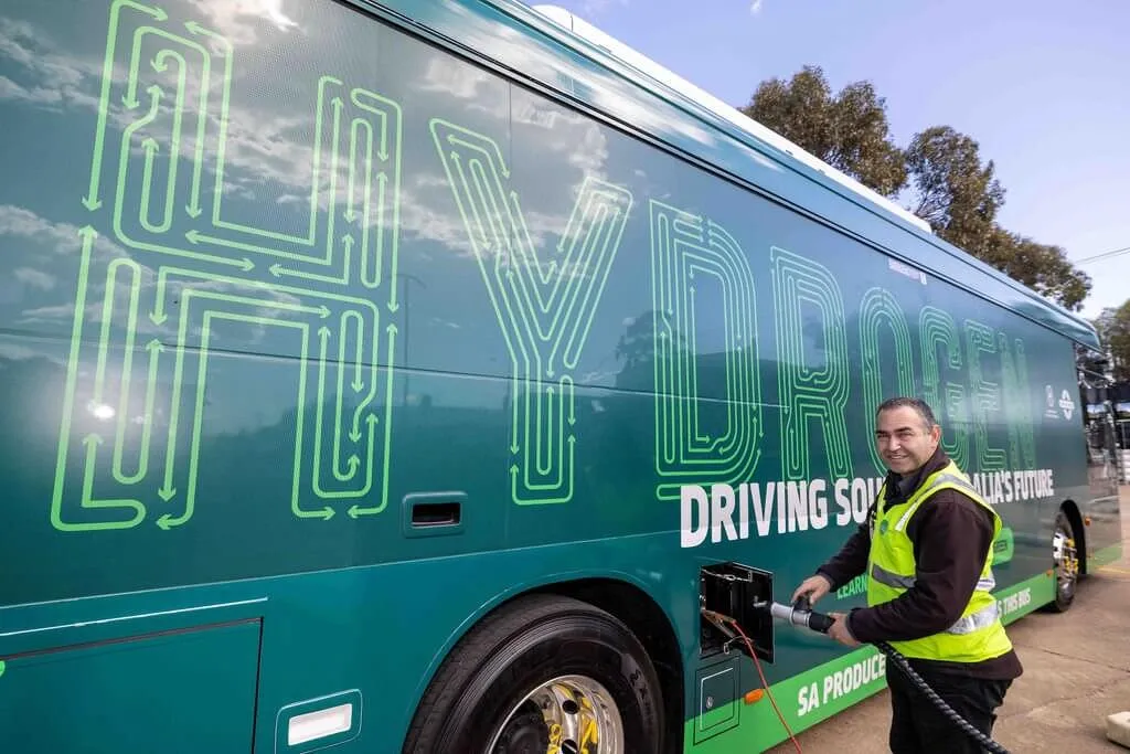 A man in a high-visibility vest refueling a green and black hydrogen bus.