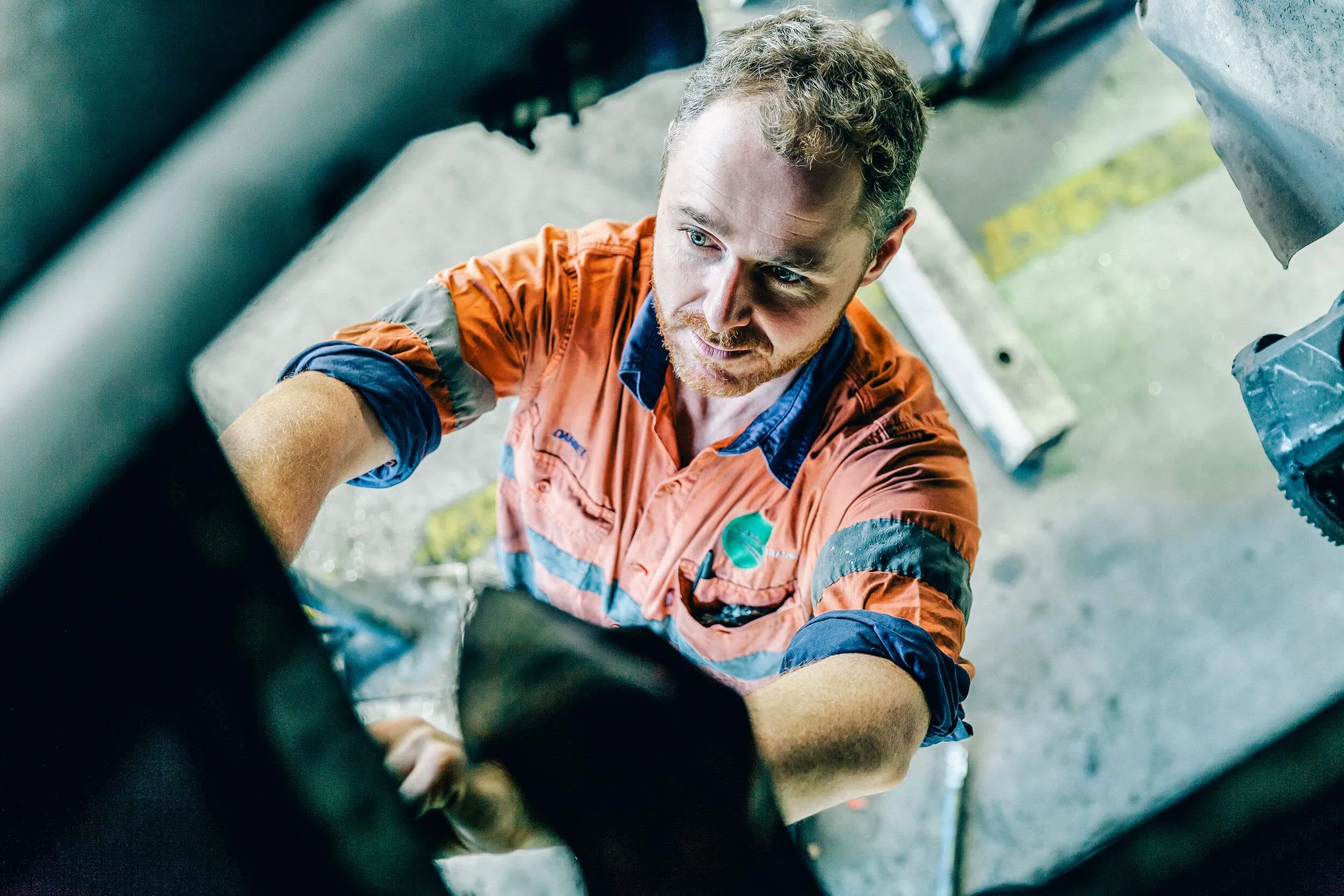 A mechanic in an orange uniform works on a vehicle, focusing intently while reaching up to make adjustments in an industrial workshop environment.