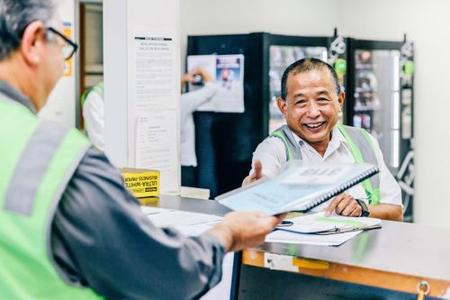 Two staff members in green safety vests interact at a counter in a workplace, one handing a document to the other who is smiling.
