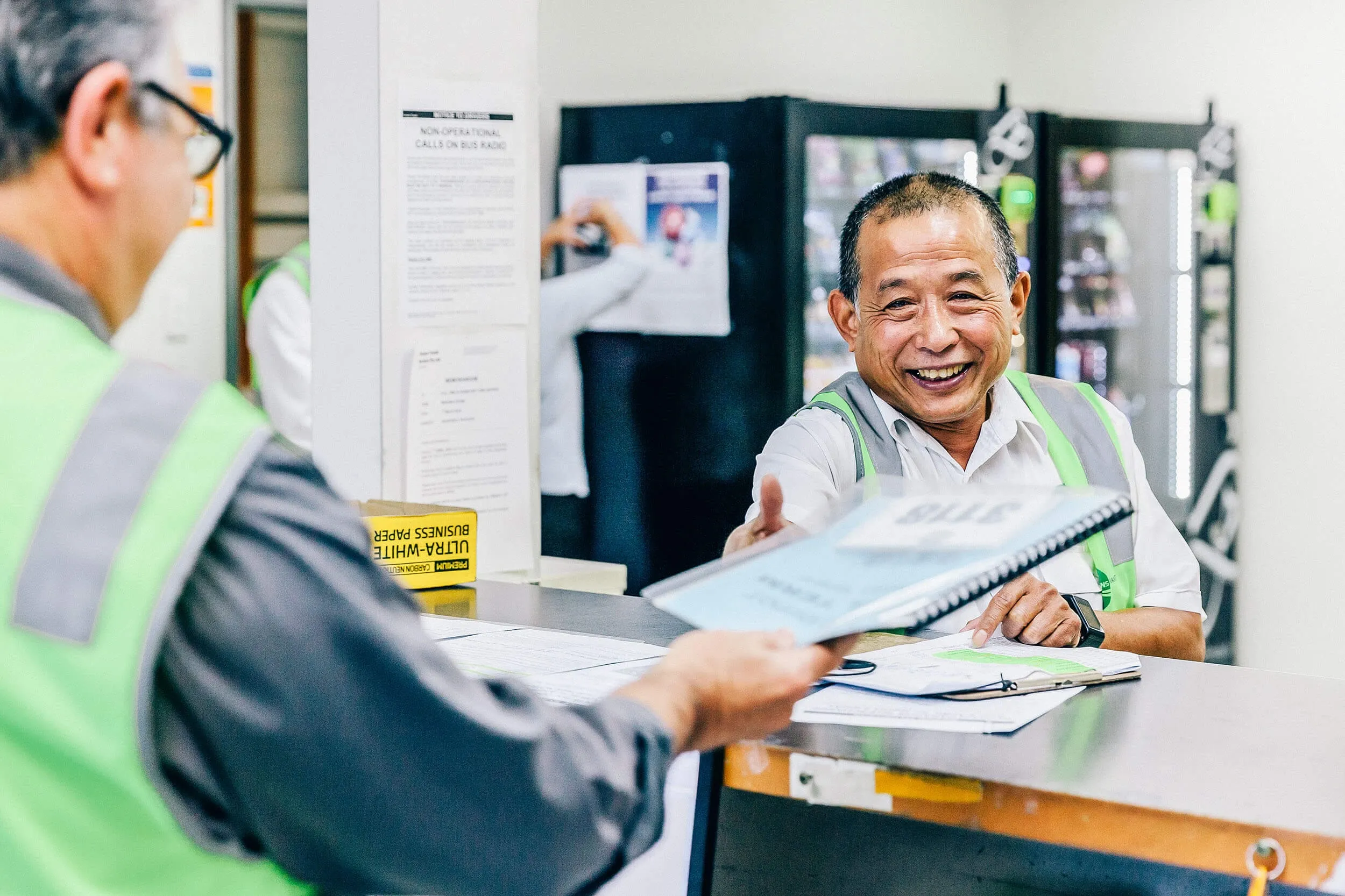 Two staff members in green safety vests interact at a counter in a workplace, one handing a document to the other who is smiling.