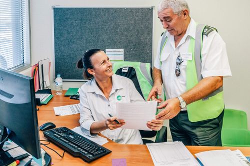 A woman seated at a desk with paperwork and a computer smiles at a standing colleague wearing a green safety vest as they discuss documents in an office setting.