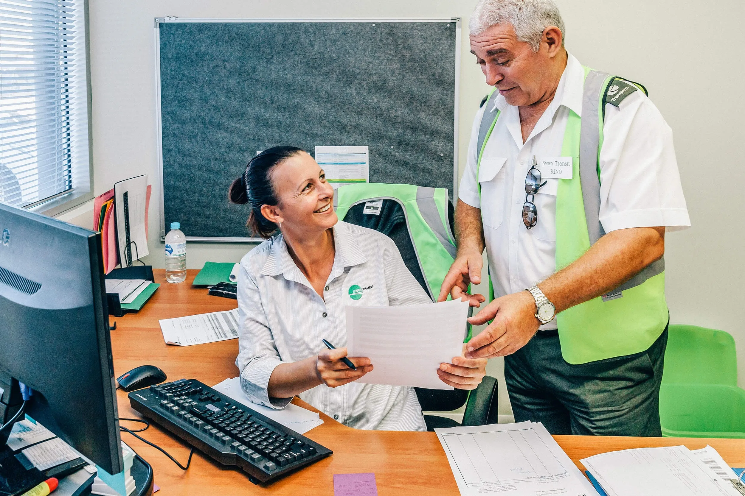 A woman seated at a desk with paperwork and a computer smiles at a standing colleague wearing a green safety vest as they discuss documents in an office setting.