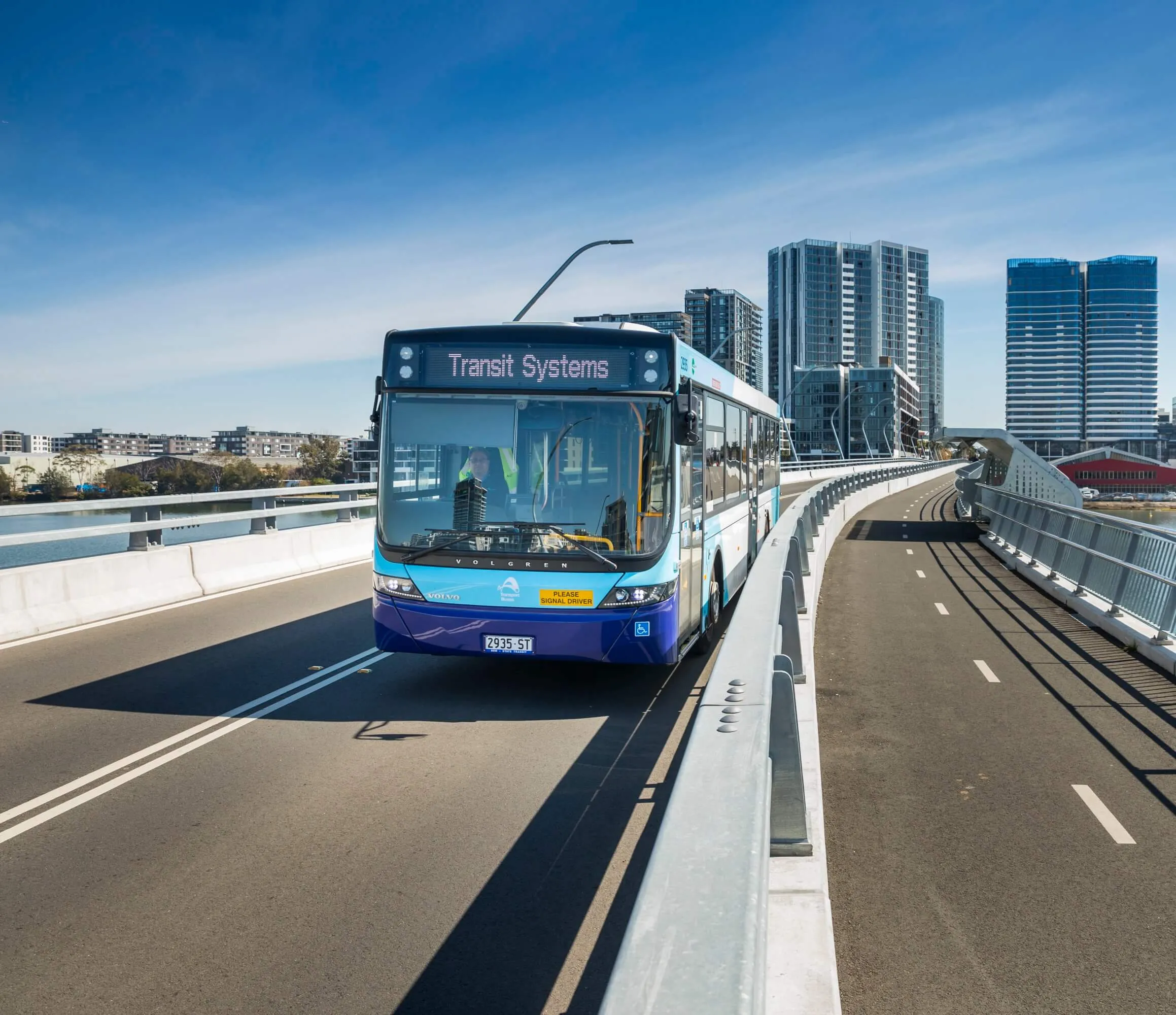 A blue Transit Systems city bus travels on a modern bridge with tall buildings in the background under a clear blue sky.
