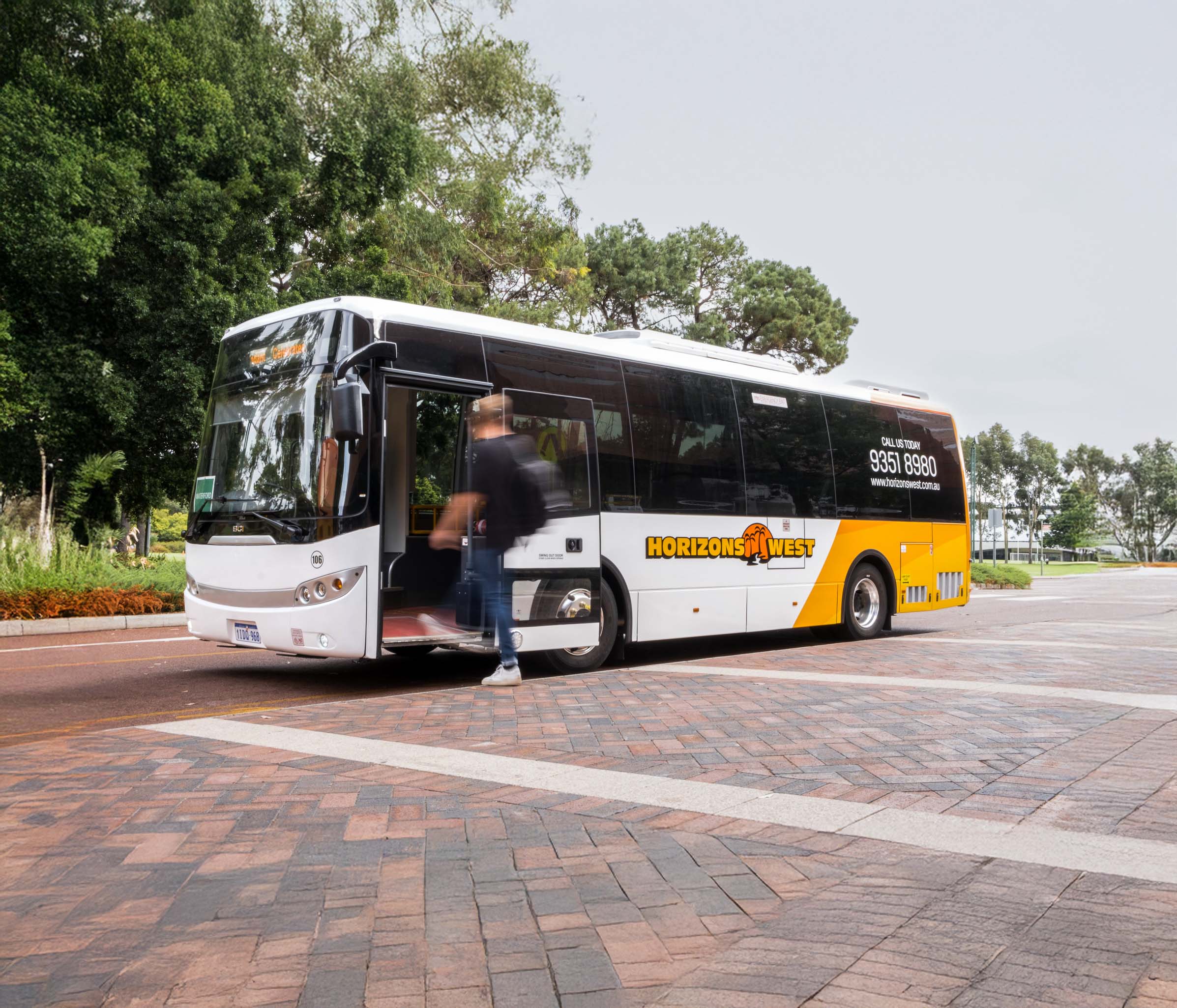 A Horizons West charter bus is parked on a brick street while a passenger boards with trees in the background.