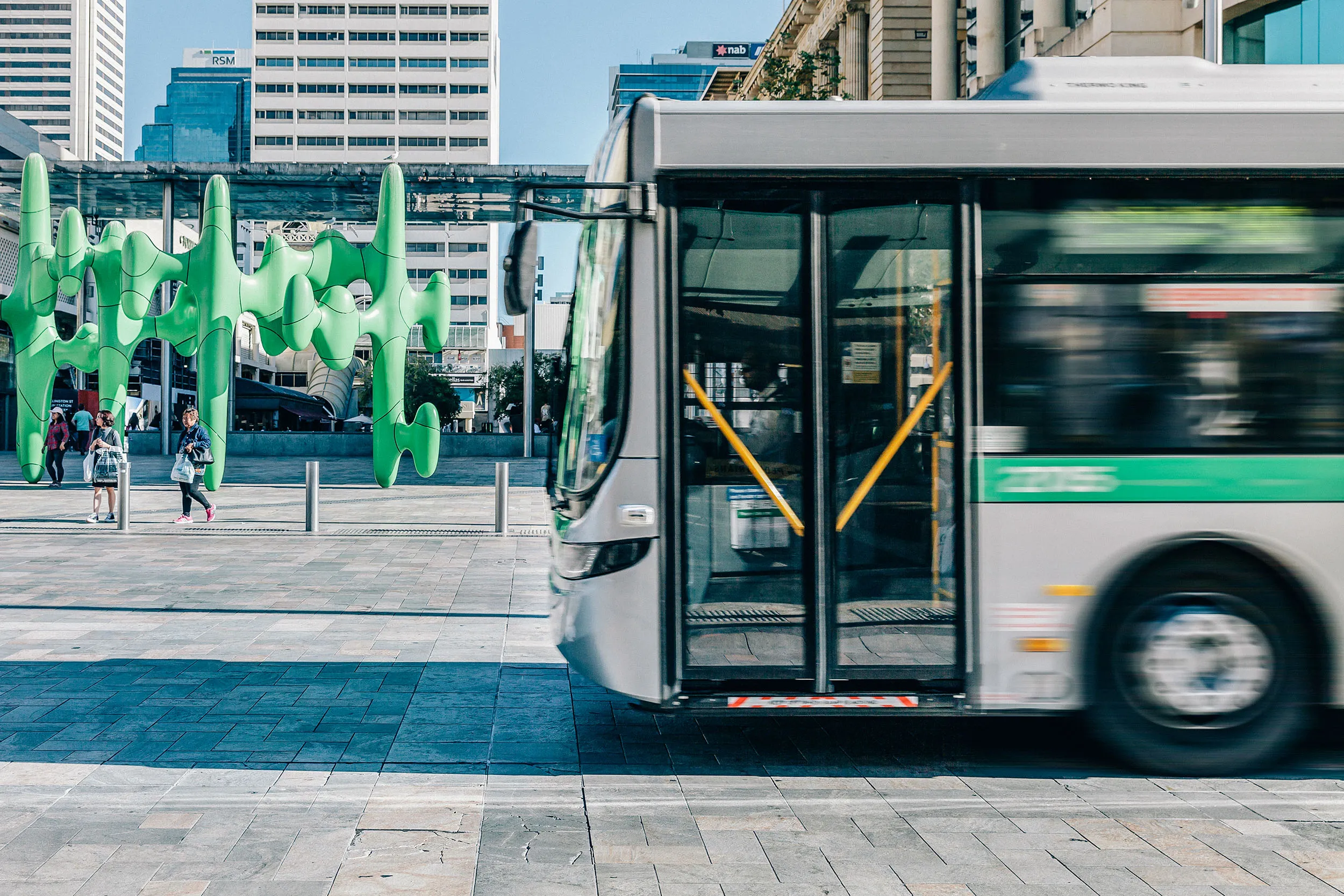 A city bus passes in front of modern green public art sculptures and tall office buildings in a downtown pedestrian plaza.