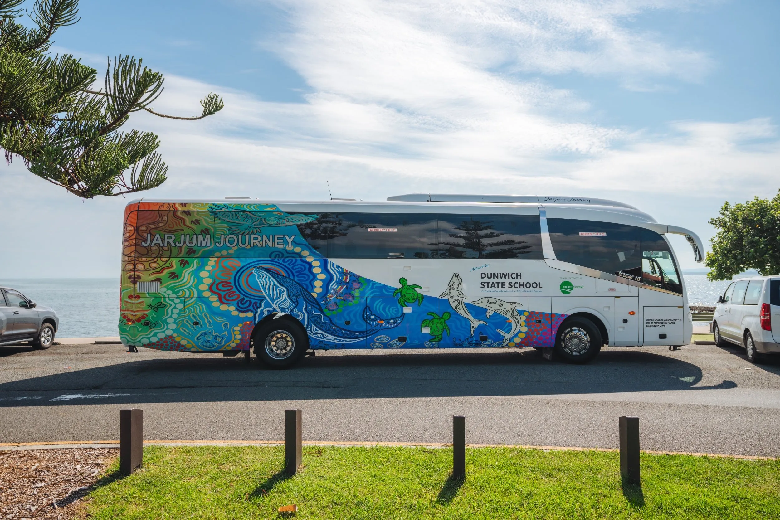 A Dunwich State School Jarjum Journey bus decorated with colorful Aboriginal artwork is parked near the ocean under a clear sky.