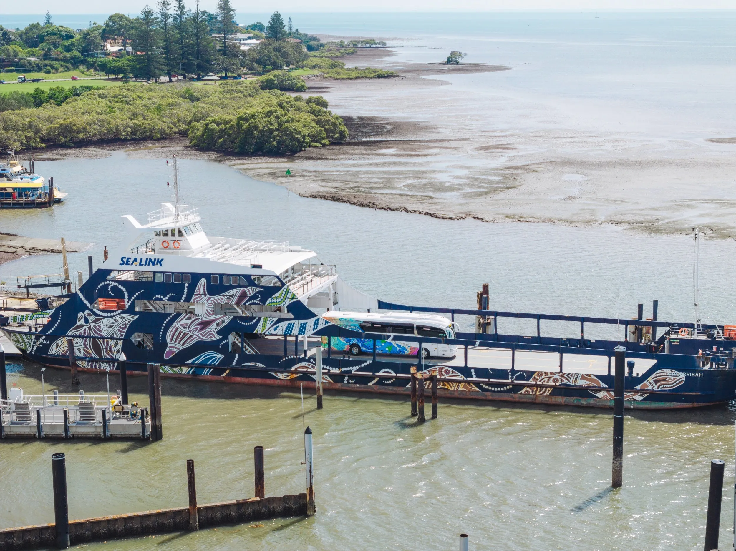 A SeaLink ferry with vibrant Aboriginal artwork docked at a terminal, carrying a bus on its deck, with a coastal landscape and mangroves in the background.