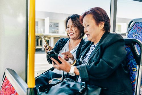 Two women sitting together on a bus, smiling and looking at their smartphones while sharing something on the screens.