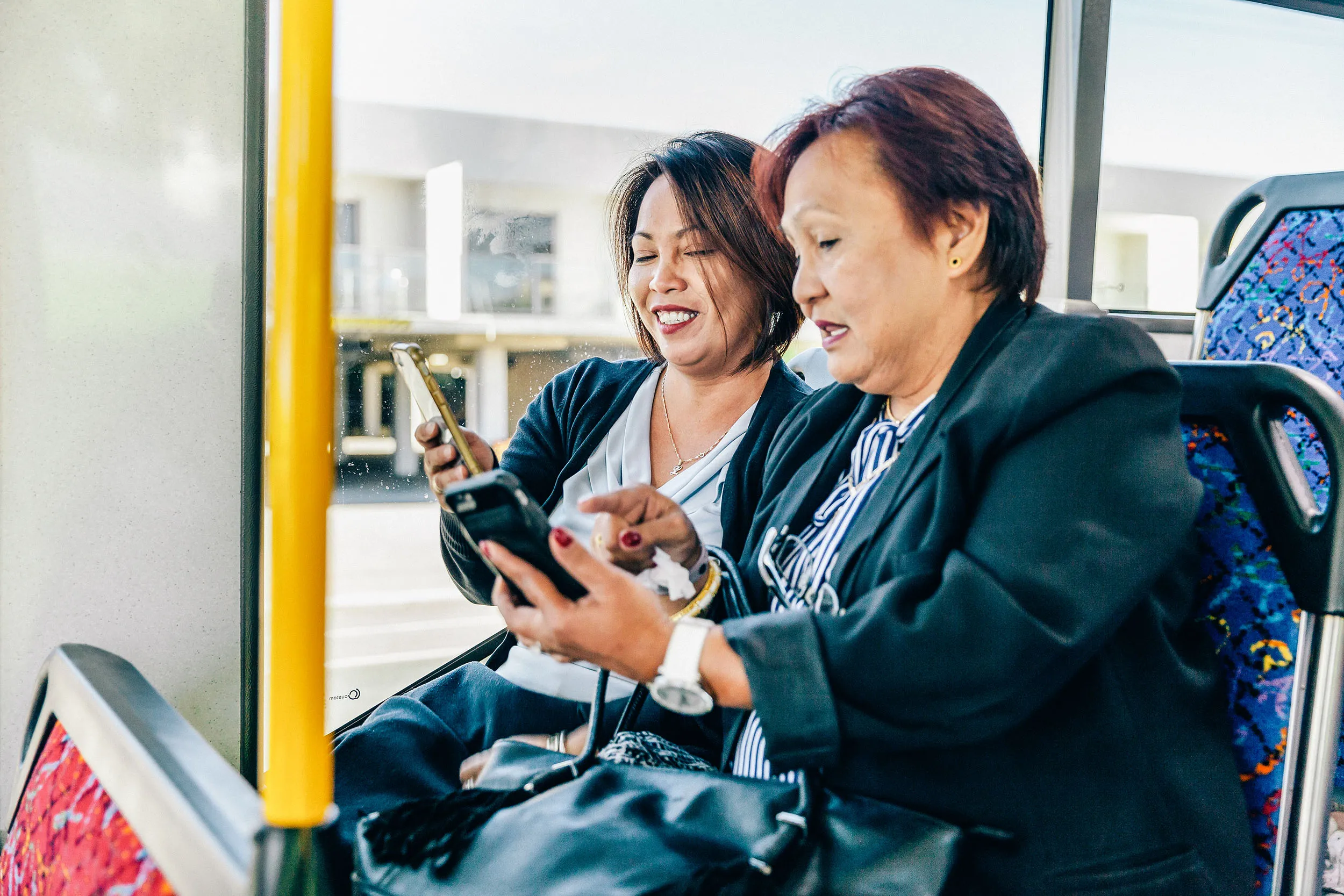 Two women sitting together on a bus, smiling and looking at their smartphones while sharing something on the screens.