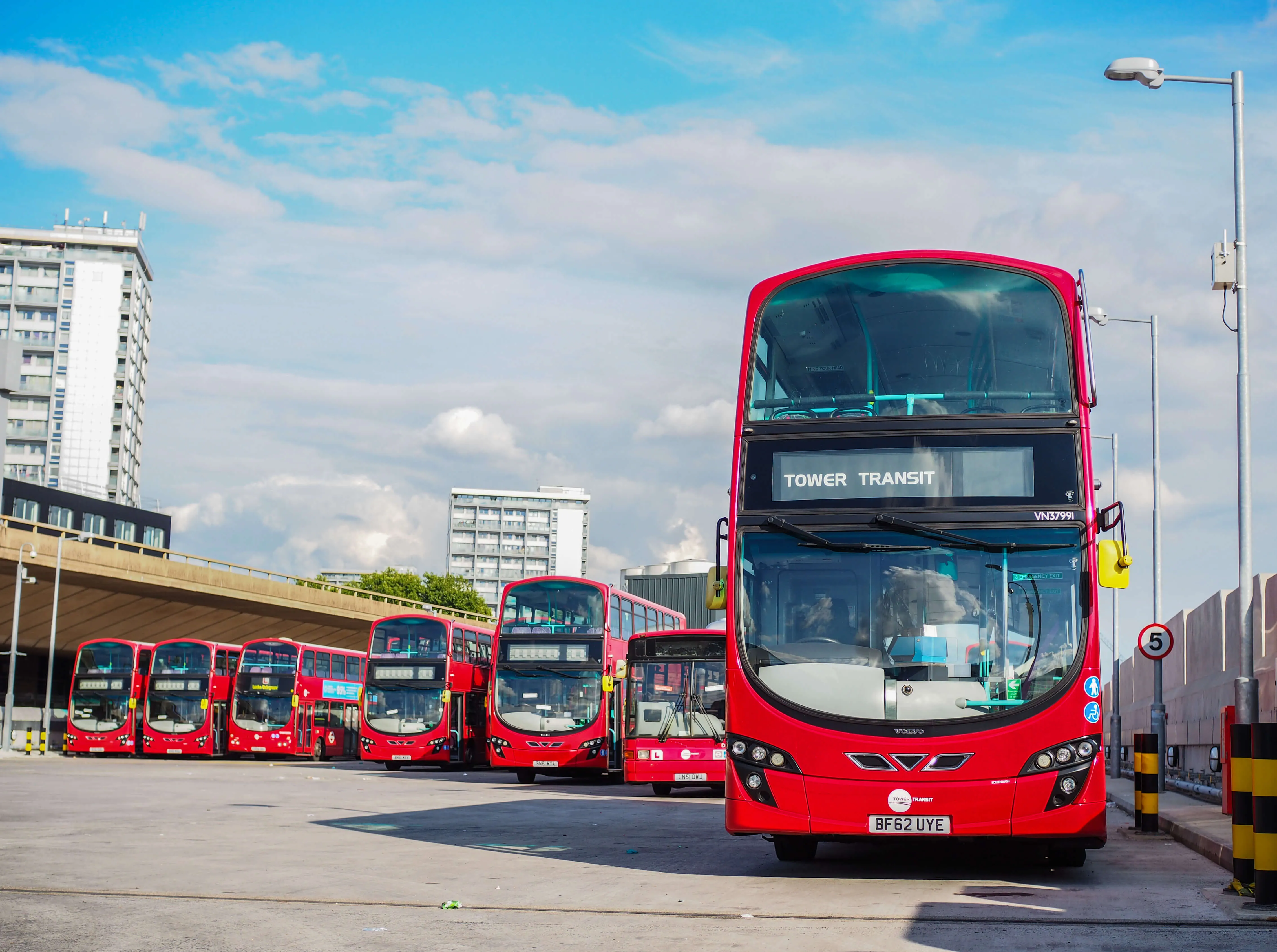 A fleet of red double-decker buses parked at a Tower Transit bus depot in London with tall apartment buildings in the background.