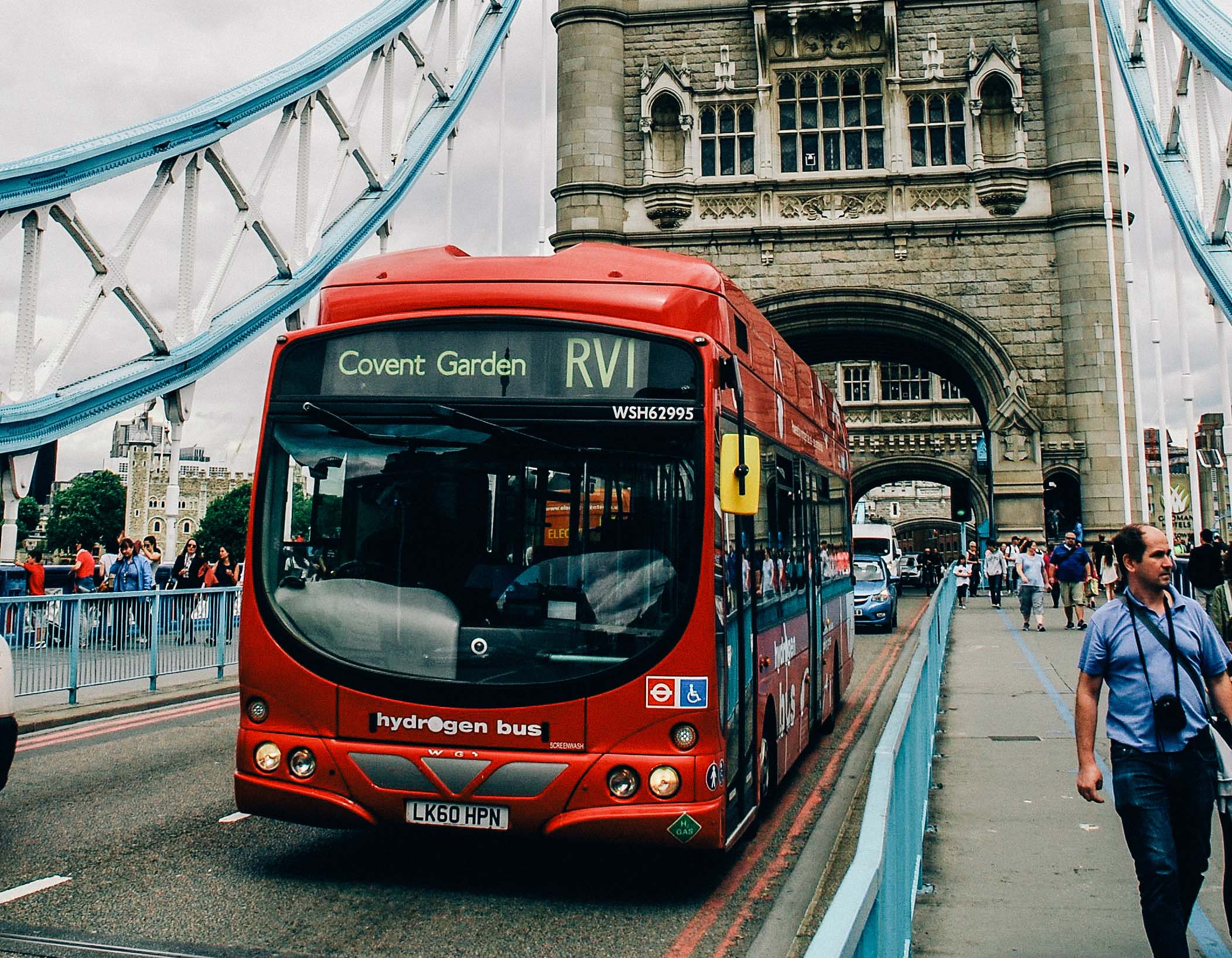 A red hydrogen bus on route RV1 to Covent Garden crossing Tower Bridge in London with people walking and historic architecture in the background.