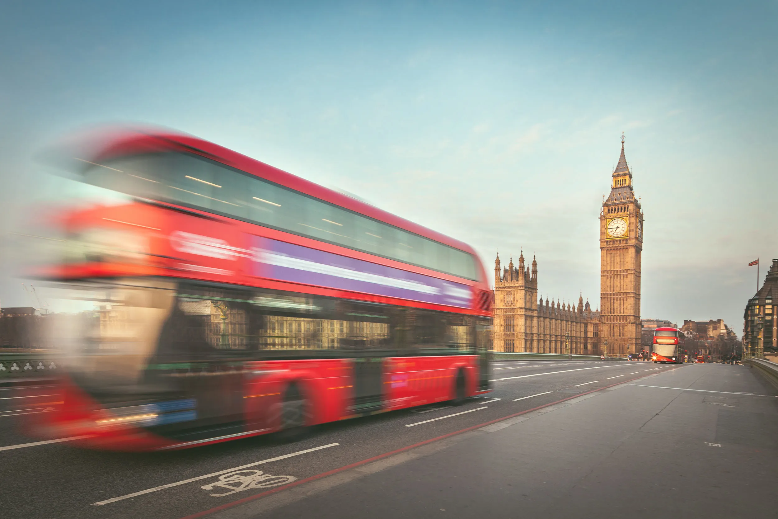 A red double-decker bus in motion crossing Westminster Bridge in London with the Houses of Parliament and Big Ben in the background at sunrise.