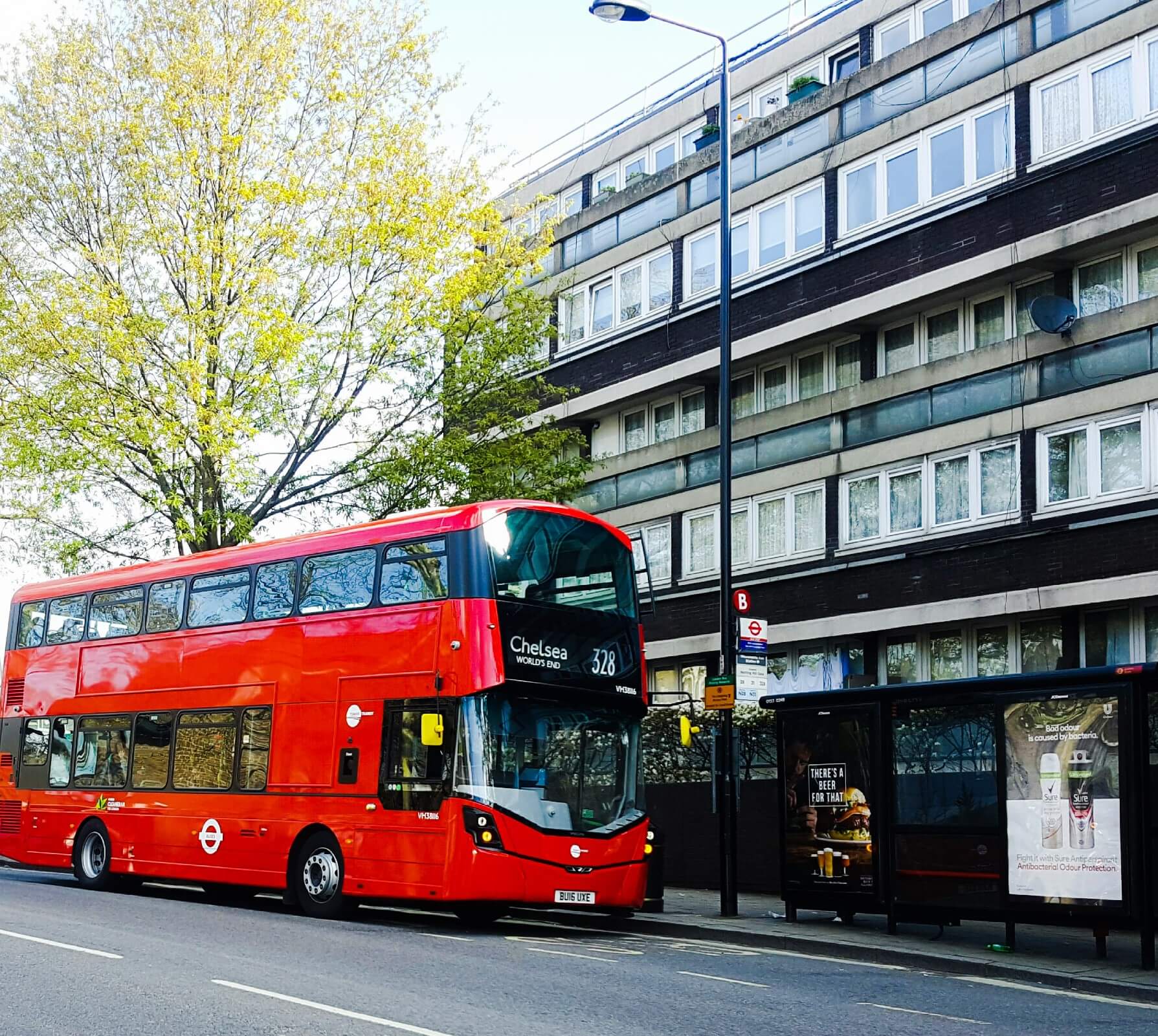 A red double-decker London bus on route 328 to Chelsea, parked at a city bus stop beside a modern apartment building and a bus shelter.