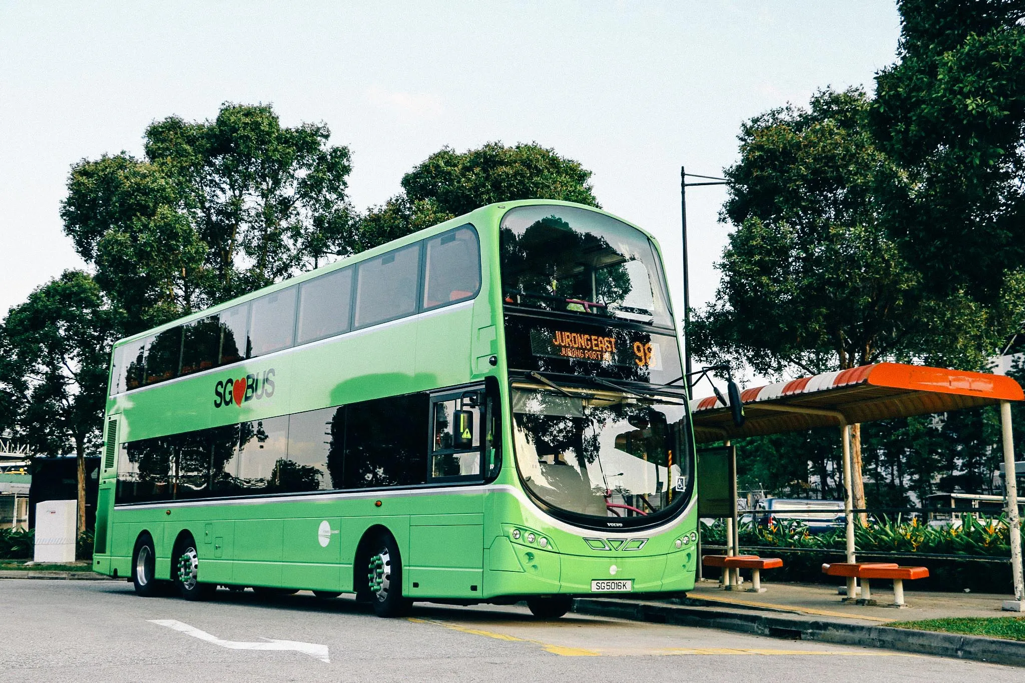 A bright green SG Bus double-decker bus on route 98 to Jurong East, parked at a bus stop in Singapore with trees and benches in the background.
