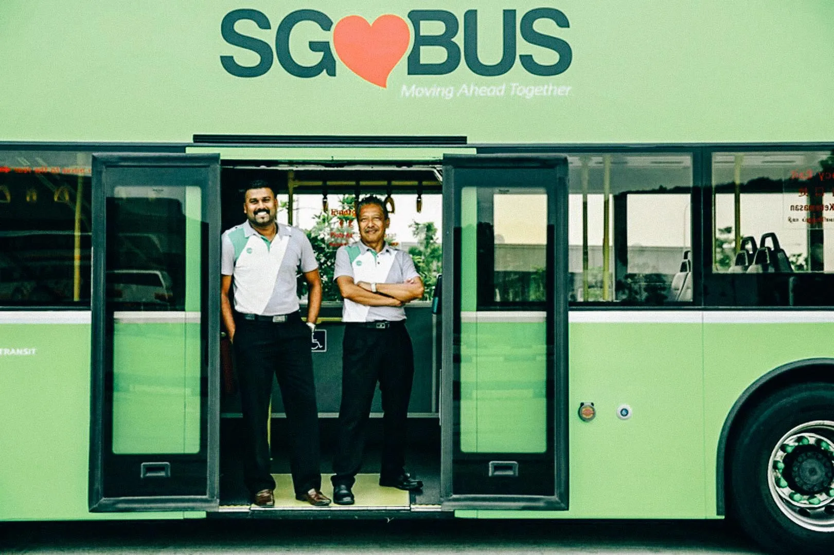 Two smiling SG Bus drivers standing at the open doors of a bright green SG Bus in Singapore, showcasing friendly public transport staff.