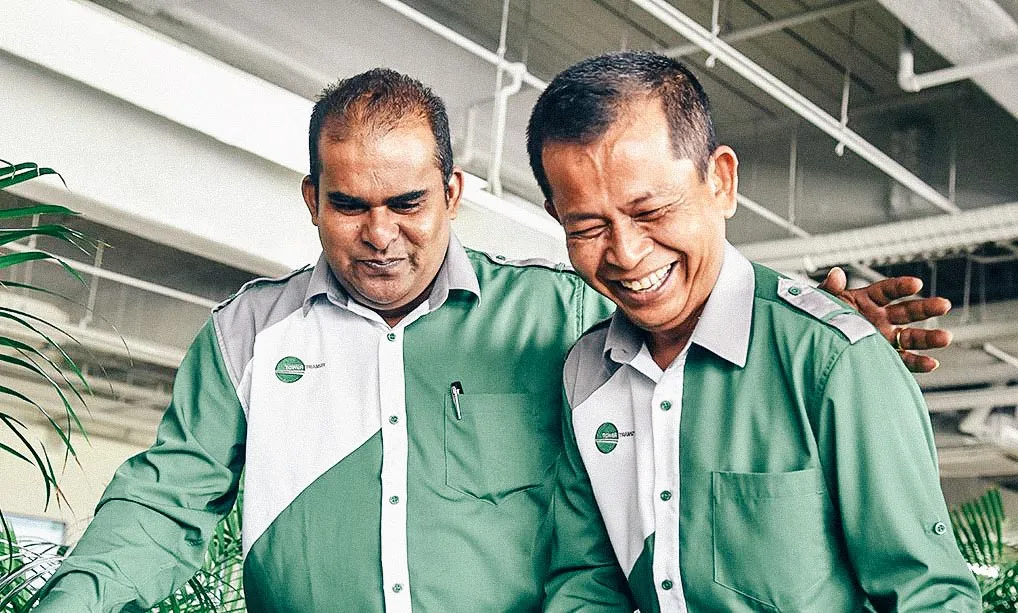 Two bus drivers in green uniforms from Tower Transit smiling and sharing a friendly moment at work.