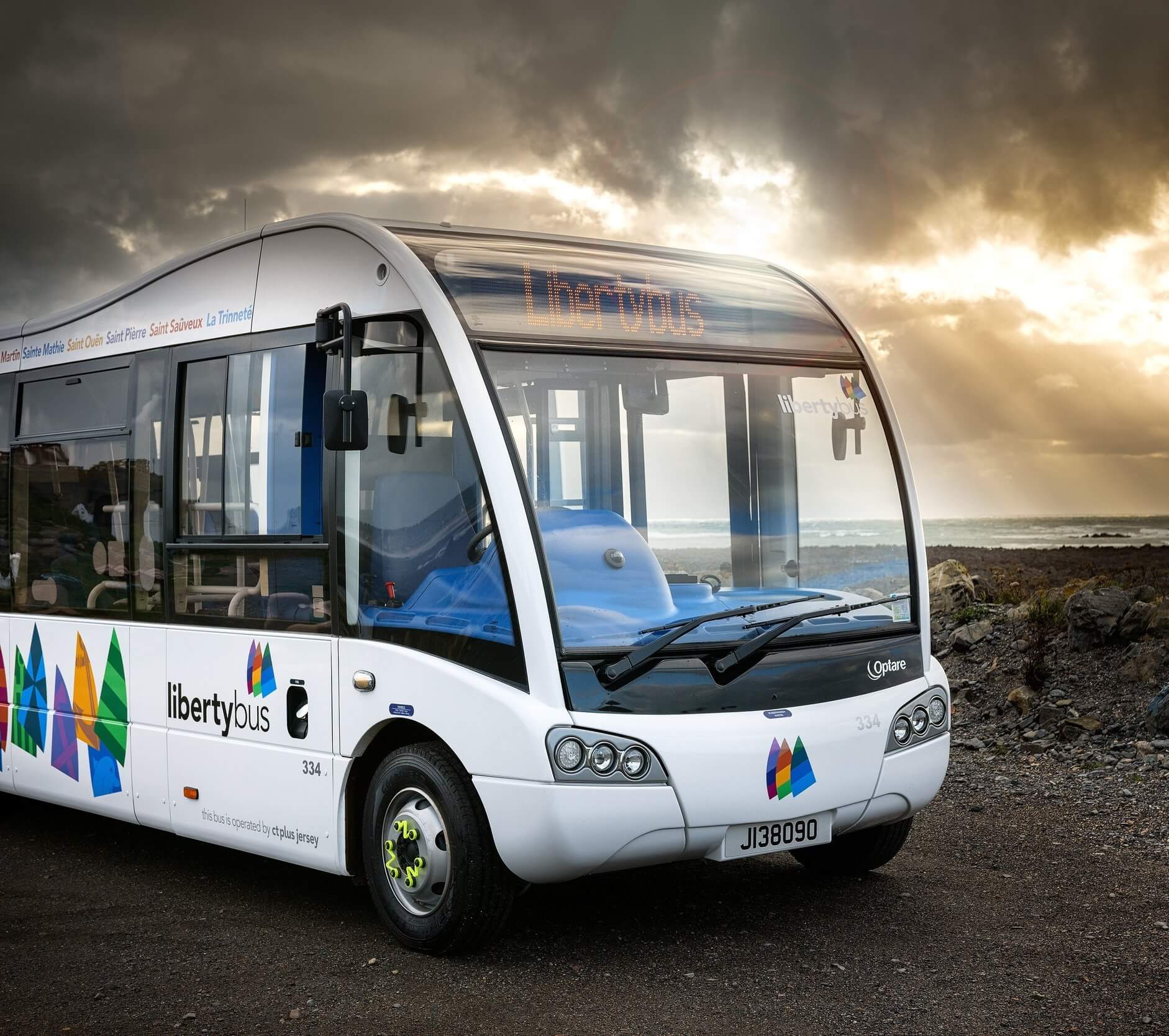 A LibertyBus parked by the coast under a dramatic cloudy sky with sunlight streaming through, showcasing its modern design and scenic location.