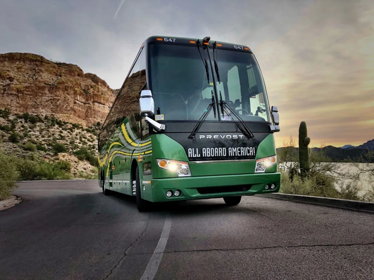 A green Prevost coach bus with "ALL ABOARD AMERICA!" branding, parked on a winding road in a desert landscape with mountains and cacti at sunset.