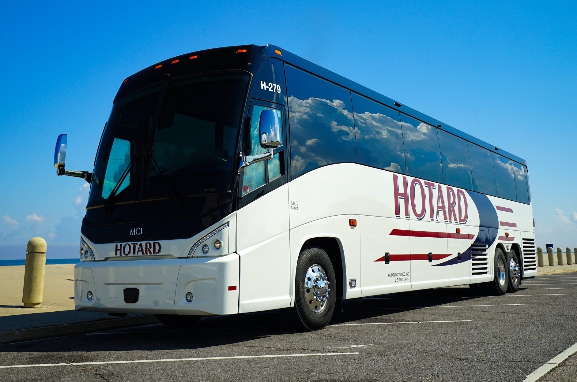 A white MCI coach bus with "HOTARD" branding parked near the ocean under a clear blue sky.
