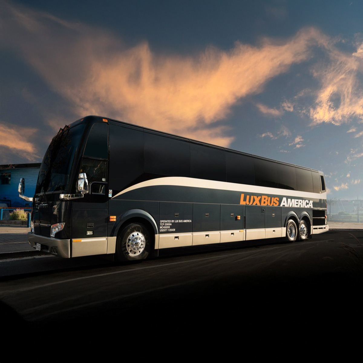 A black and silver Lux Bus America coach bus parked on a street at sunset, with a dramatic sky in the background.