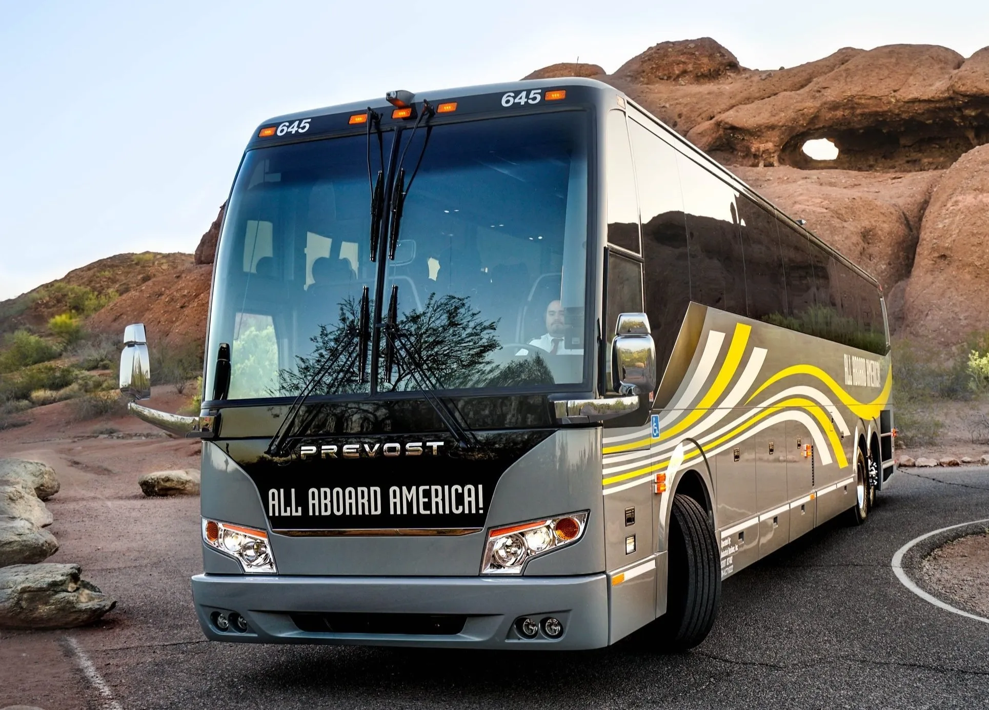 A grey and black Prevost coach bus with "ALL ABOARD AMERICA!" branding, parked on a winding road next to red rock formations.