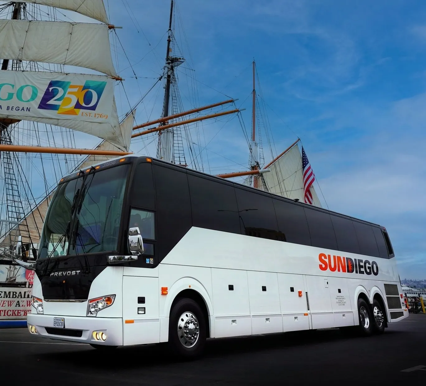 A white Prevost coach bus with "San Diego" branding parked in front of a historic sailing ship with large sails and an American flag, under a blue sky.