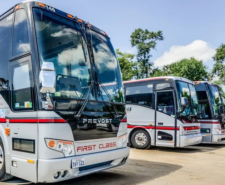 Several silver and black Prevost "First Class" coach buses parked in a row under a blue sky with trees in the background.
