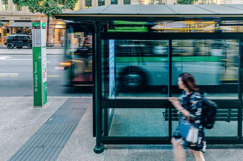 A city bus stop on St Georges Terrace with a blurred bus passing by and a woman walking past, creating a sense of motion in an urban setting.