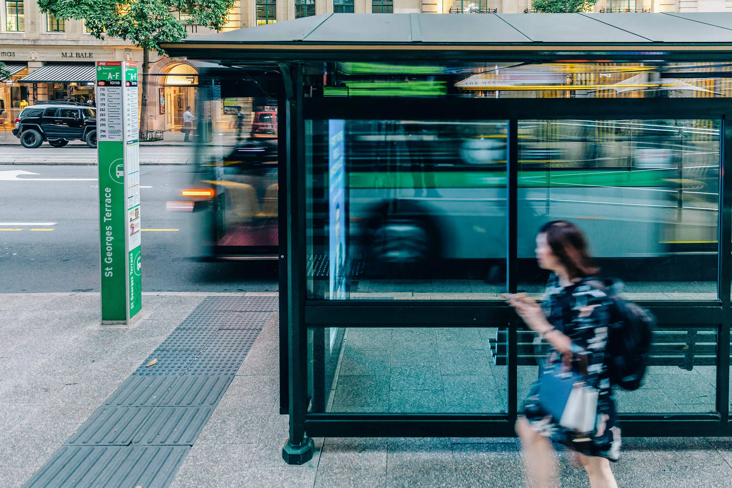 A city bus stop on St Georges Terrace with a blurred bus passing by and a woman walking past, creating a sense of motion in an urban setting.