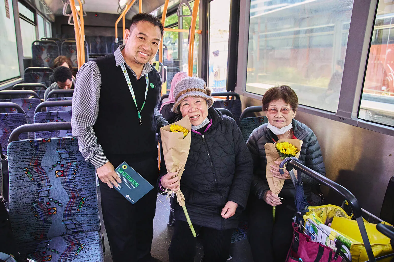 A bus driver standing and smiling with two elderly women who are seated and holding sunflowers, inside a city bus.
