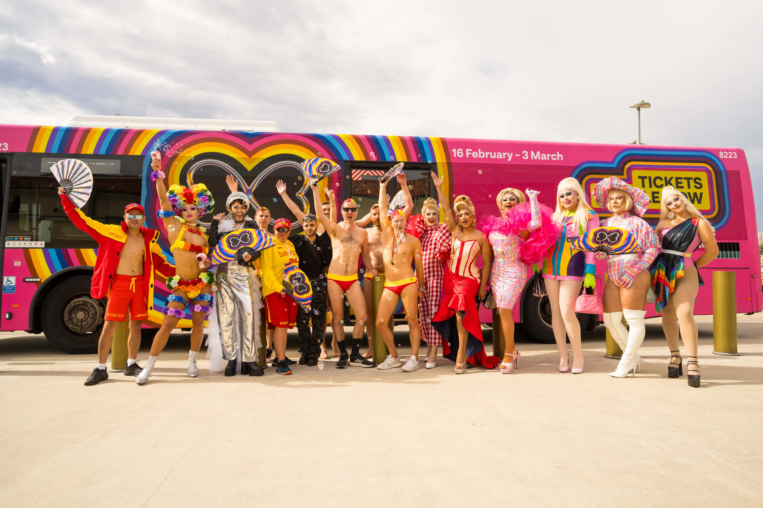 A vibrant group of people in colorful Mardi Gras costumes and surf rescue outfits posing together in front of a bright pink Transit Systems bus decorated for Sydney Mardi Gras.