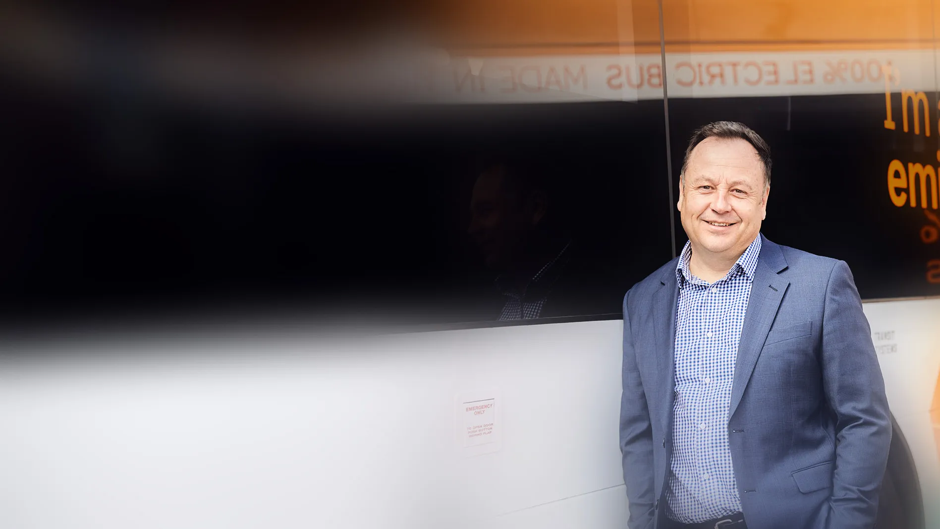 A man in a blue suit stands smiling next to a colorful bus with the words "I'm a zero emissions bus" displayed on its side.